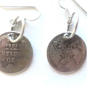May include: A pair of silver earrings featuring vintage coins. Each coin is round with embossed details and attached to a silver hook. The coins have visible text and the year 1864.