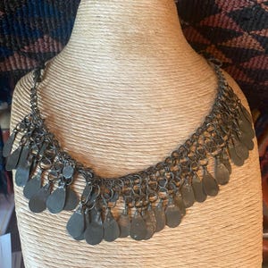 May include: A silver-toned necklace with a chain design and numerous teardrop-shaped pendants. The necklace is displayed on a tan mannequin, showcasing its intricate details and vintage aesthetic. The background features a patterned textile.