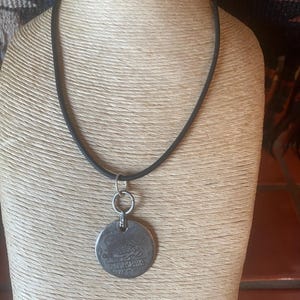 May include: A silver-colored pendant necklace with a black cord. The pendant is a round coin with etched details and text. The necklace is displayed on a mannequin.