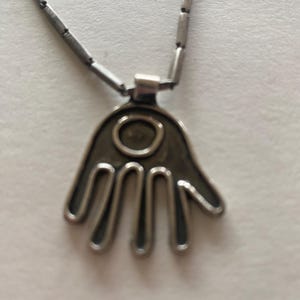 May include: Silver-toned necklace featuring a hand-shaped pendant with an oval center. The pendant has five fingers and is connected to a chain of small, rectangular links. The necklace is displayed against a white background.