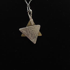 May include: A silver necklace with a pendant featuring a textured, hammered silver triangle over a gold-toned triangle. The pendant hangs from a delicate silver chain against a black background.