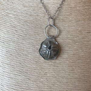 May include: A silver-toned necklace featuring a pendant with an embossed floral design. The pendant is attached to a chain necklace. The necklace is displayed against a neutral background.