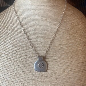 May include: A silver-colored pendant necklace. The pendant has a rounded shape with a central circular design and a small rectangular bail. The chain is a delicate link style. The necklace is displayed on a neutral-toned mannequin.