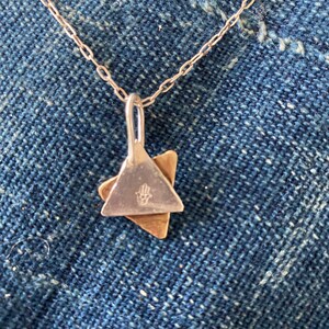 May include: Silver necklace with a pendant featuring two overlapping triangles, one silver and one gold. The silver triangle has a symbol on it. The necklace is displayed on a blue denim background.