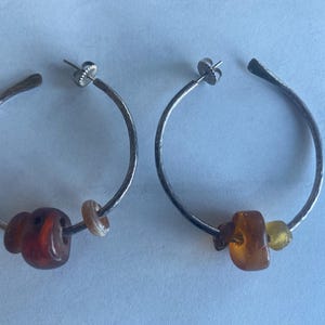May include: A pair of silver hoop earrings with hammered texture. Each earring features a combination of amber and light-colored beads. The earrings are displayed on a light blue surface.