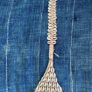 May include: A decorative item made of white seashells attached to a light brown leather strip. The seashells are arranged in a vertical line and a triangular shape. The background is a blue fabric with white stripes.
