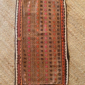 May include: Rectangular, hand-stitched textile with a repeating geometric pattern in shades of brown, orange, pink, and black. The border is a darker brown with a scalloped edge. The textile is displayed on a woven, tan surface.