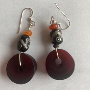 May include: A pair of handcrafted earrings featuring dark red, circular beads with a silver wire and hook. The earrings also include black beads with silver designs and small orange beads.