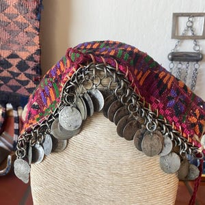 May include: A colorful, patterned textile head covering with silver coin accents. The fabric features a mix of pink, orange, green, and purple hues. Silver chain and coin embellishments hang from the edge of the head covering.