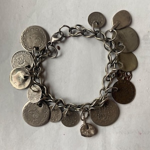 May include: Silver-toned charm bracelet featuring a variety of antique coins. The bracelet has a chain link design with a lobster clasp closure. The coins vary in size and design, adding a unique, vintage aesthetic to the jewelry.