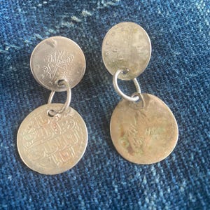 May include: A pair of silver-toned dangle earrings. Each earring features two circular coin-like pendants connected by a small ring. The coins have intricate designs and text. The earrings rest on a blue denim surface.