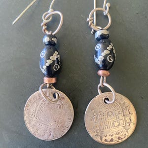 May include: A pair of handcrafted earrings featuring silver-tone metal hooks and coin-shaped pendants. Each earring has a black bead with white floral designs and a copper-colored connector. The coin pendants display an embossed architectural design.