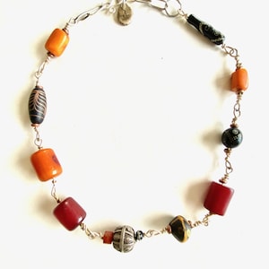 May include: A handcrafted necklace featuring a variety of beads in shades of orange, red, black, and yellow. The beads are strung together with silver-colored wire and chain, creating a unique and colorful accessory. The necklace is displayed on a white background.