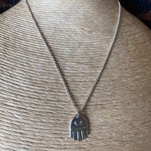May include: A silver-toned necklace featuring a pendant shaped like a hand with an eye design. The pendant hangs from a delicate chain. The necklace is displayed against a textured, neutral-toned background.