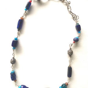 May include: A handcrafted necklace featuring a variety of beads in shades of blue, brown, and silver. The necklace includes dark blue rectangular beads, small round beads, and decorative silver beads, strung on a silver chain.