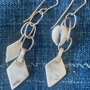 May include: A pair of silver earrings with a diamond-shaped pendant. One earring features two small diamond pendants, while the other has a cowrie shell. The earrings are connected by a series of small silver rings and a hook.