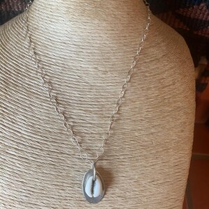 May include: A silver necklace featuring a cowrie shell pendant. The pendant is an oval shape with a silver border and a white and beige shell. The chain is a linked design. The necklace is displayed on a mannequin.