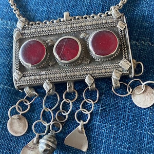 May include: A silver-toned necklace with a rectangular pendant featuring three red circular stones. The pendant is intricately detailed with decorative patterns and small dangling charms, including round and triangular shapes, and a small bell.