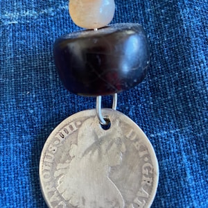 May include: A handcrafted necklace featuring a silver coin pendant dated 1794, suspended below a dark, rounded bead and a light-colored stone. The necklace is strung on a black cord, creating a unique, vintage-inspired accessory.
