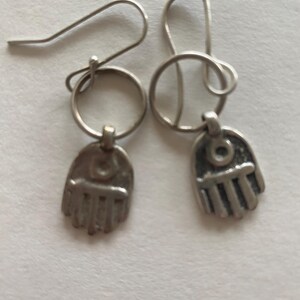 May include: A pair of silver-toned earrings. Each earring features a small, open hand charm with detailed fingers and a central eye design. The earrings have a simple hook closure.