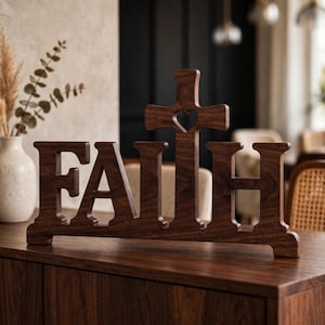 Op de afbeelding: Een donkerbruin houten decoratief bord met het woord FAITH in blokletters, waarbij de letter T is vervangen door een kruisvorm met een hartvormige uitsparing in het midden, rustend op een platte voet voor extra stabiliteit.