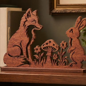 May include: A brown wooden cutout decoration featuring a fox sitting on the left and a rabbit standing on the right, separated by a central mushroom and small flowers. The entire piece is mounted on a flat rectangular wooden base.