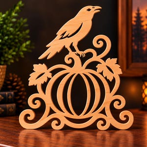 Pode incluir: Um recorte de madeira de cor clara apresentando um corvo pousado sobre uma abóbora. O design inclui volutas decorativas na base e duas folhas ladeando a abóbora. A peça inteira é feita a partir de uma única folha de madeira plana.