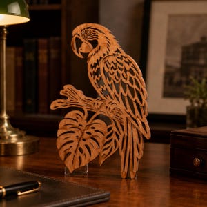 May include: A laser-cut wooden parrot sculpture perched on a branch with a large monstera leaf below. The artwork features intricate layered details and a natural wood grain finish, standing upright on a clear acrylic base for display.