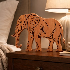 May include: A light brown wooden elephant figurine featuring dark, laser-cut lines that define the animal's ears, trunk, legs, and body contours. The cutout design creates a silhouette of an elephant standing in profile with its trunk curved.