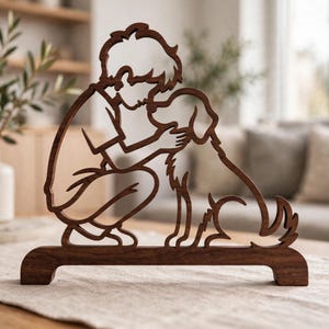 May include: A dark brown wooden cutout sculpture featuring the silhouette of a person crouching down to pet a dog. The design is connected by a solid horizontal base, creating a continuous outline of the figures in a warm, natural wood tone.