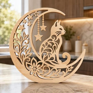 May include: A light-coloured wooden cutout decoration shaped like a crescent moon. The design features an intricate filigree pattern of a cat sitting on the moon, surrounded by floral motifs, leaves, and hanging star shapes within the moon's curve.