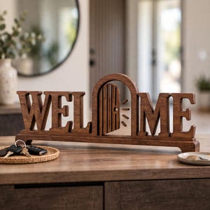 Könnte beinhalten: Ein dunkelbraunes Holzschild mit dem Wort WELCOME in Blockbuchstaben, bei dem der Buchstabe O durch ein Bogentür-Design mit einer kleinen Tür und strahlenförmigen Linien ersetzt ist. Das Schild steht auf einem flachen Sockel mit natürlicher Optik.