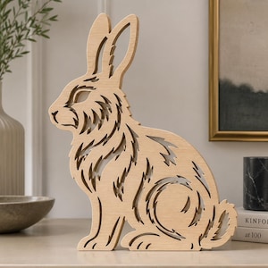 May include: A light-colored wooden cutout of a rabbit in a sitting position. The figure features intricate, stylized cut-out patterns throughout its body, including the ears, fur, and tail, creating a layered, open-work design with a smooth finish.