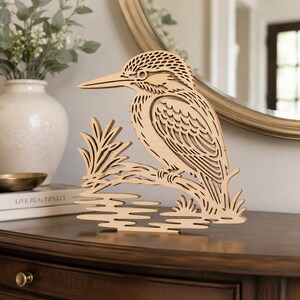 Könnte beinhalten: Eine hellfarbene, lasergeschnittene Holzskulptur, die einen Eisvogel auf einem Ast über stilisiertem Wasser darstellt. Das Design zeichnet sich durch komplexe, geschichtete Ausschnitte aus, die das Gefieder, den Schnabel und die natürlichen Elemente definieren.