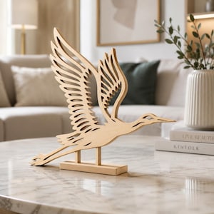 May include: A light-coloured wooden sculpture of a bird in flight with wings spread wide. The bird is depicted in a stylised, cutout design with intricate feather details along the wings. It is mounted on a rectangular wooden base with two supports.