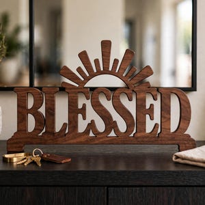 May include: A dark brown wooden sign featuring the word BLESSED in a bold, sans-serif font. The top of the sign is decorated with a stylised sunburst design consisting of radiating rays. The sign is freestanding with a flat base.