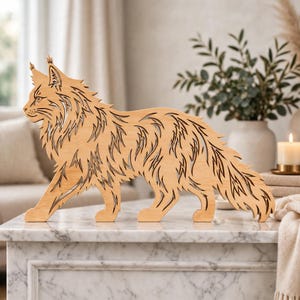May include: A light brown wooden cutout of a Maine Coon cat standing in profile. The figure features intricate laser-cut details depicting the cat's long, shaggy fur, pointed ears with tufts, and a long, bushy tail, all rendered in a flat, silhouette style.
