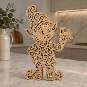 May include: A light brown wooden cutout of an elf figure featuring intricate scrollwork patterns throughout the body, hat, and shoes. The elf is depicted smiling and holding a small toy train in one hand, showcasing detailed laser-cut craftsmanship.