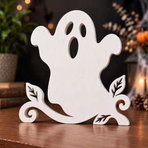 May include: A white wooden cutout of a ghost with two oval eyes and an oval mouth, featuring a decorative base with swirling vine-like branches and small leaf shapes extending from the bottom on both the left and right sides of the figure.