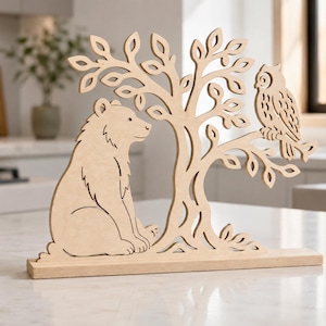 May include: A light-colored wooden cutout decoration featuring a bear sitting at the base of a tree with an owl perched on a branch. The entire piece is mounted on a flat rectangular wooden base, showcasing intricate laser-cut details.