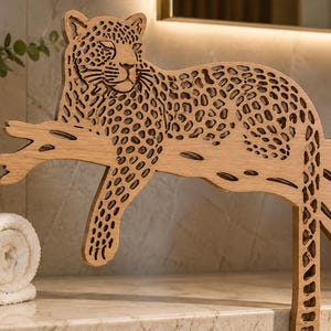 Può includere: Una figura di leopardo in legno tagliata al laser che riposa su un ramo. Il leopardo presenta un dettagliato motivo traforato che rappresenta le sue macchie, con una finitura naturale in venature del legno marrone chiaro. Il design mostra l'animale.