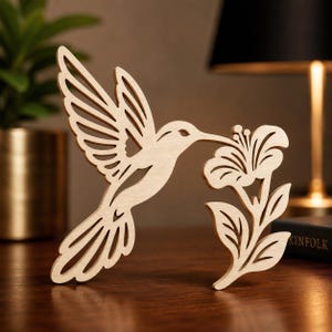 May include: A light-colored wooden cutout featuring a hummingbird in flight with its beak touching a flower. The design is composed of clean, open-work lines that define the bird's wings, body, and tail, alongside a stylized floral stem.