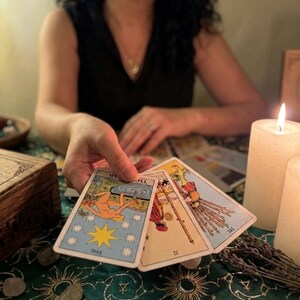 Pode incluir: Tr&ecirc;s cartas de tar&ocirc; seguradas em uma m&atilde;o, incluindo a carta A Estrela com uma figura ajoelhada, a carta Dois de Paus mostrando uma pessoa segurando um globo, e o Dez de Paus retratando uma figura carregando um feixe de dez bast&otilde;es.
