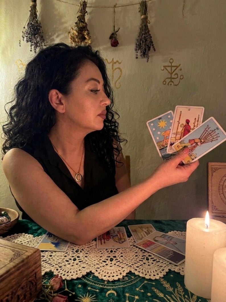 Pode incluir: Um conjunto de cartas de tarot com A Estrela, Dois de Paus e Dez de Paus, seguradas numa m&atilde;o. As cartas exibem ilustra&ccedil;&otilde;es coloridas de figuras e s&iacute;mbolos. Outras cartas de tarot est&atilde;o espalhadas numa superf&iacute;cie verde com uma renda branca.