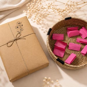May include: A rectangular gift box wrapped in brown paper with a black floral line drawing and tied with thin twine, positioned next to a round woven seagrass basket containing several small, folded bright pink paper slips.