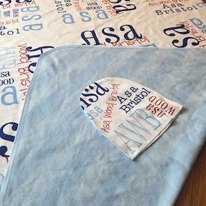 May include: A blue and white baby blanket with the name "Asa" and "Bristol" printed on it. The blanket is folded in half and a white hat with the same name is on top of it.