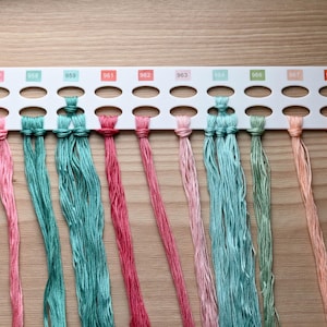 May include: A white plastic embroidery floss organiser card featuring ten numbered slots labelled 957, 958, 959, 961, 962, 963, 964, 966, 967, and 970. Strands of embroidery thread in shades of pink, teal, green, peach, and orange are looped through the slots.
