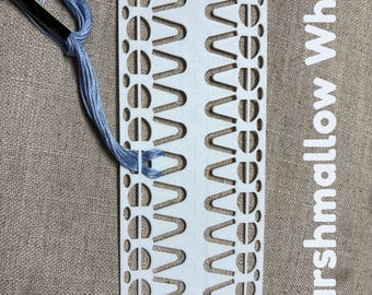 以下が含まれることがあります： 複数のU字型スロットと円形の穴が開いた白いプラスチック製の刺繍糸整理用カード。1つのスロットに水色の刺繍糸が通されており、側面にはMarshmallow Whiteという文字が印刷されています。