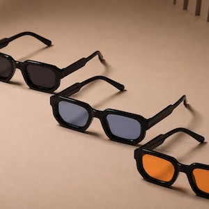 Peut inclure: Trois paires de lunettes de soleil avec des montures noires épaisses disposées en diagonale. La paire du haut a des verres gris foncé, celle du milieu des verres bleu clair et celle du bas des verres orange vif. Chaque paire est carrée.