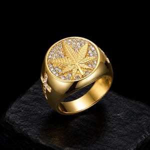 Peut inclure: Une bague dorée présentant une face circulaire avec un motif de feuille de cannabis en relief au centre. Le fond de la face circulaire est incrusté de nombreuses petites pierres claires et scintillantes. Les côtés de l'anneau portent une croix.